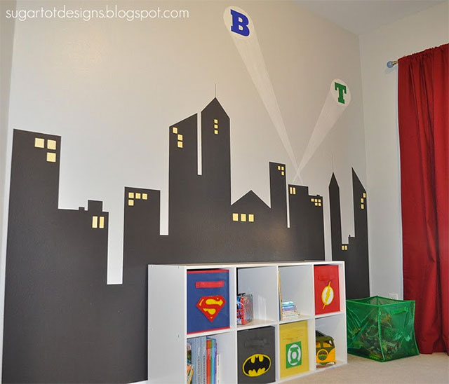 Pared pintada con la silueta de una ciudad de noche con dos halos de luz y unas iniciales, a lo Batman. Mueble modular de ocho secciones, apoyado contra la pared pintada, algunas secciones cerradas y en el frente de cada una de las puertas del mueble, los símbolos de Batman, Superman, Linterna Verde y Flash.