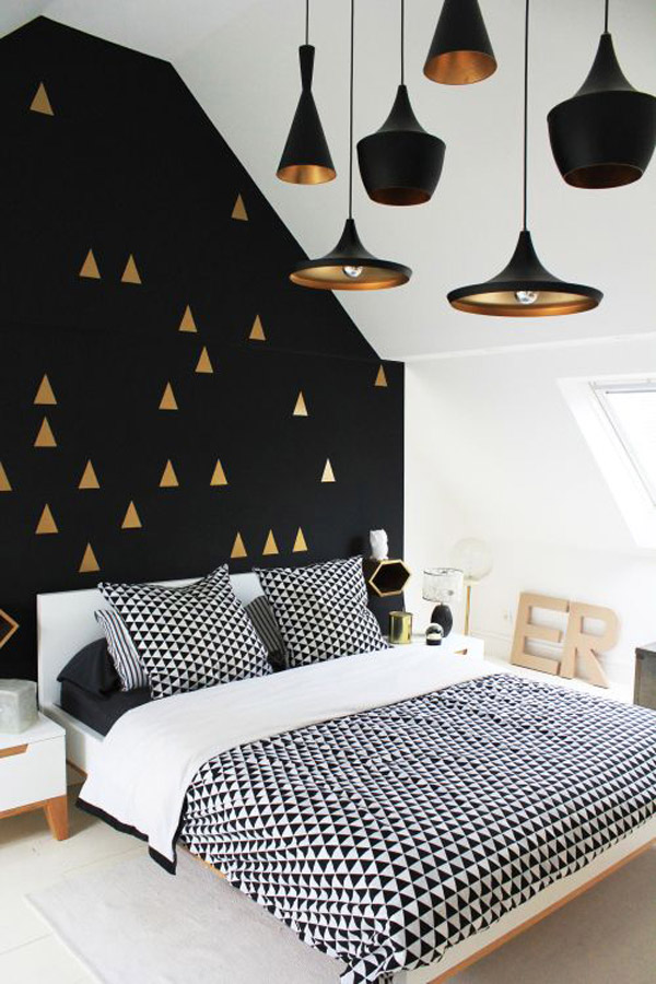 Dormitorio con pared de la cabecera pintada negra con triangulos dorados. La cama tiene un cubrecama, con sus respectivos cojines, de triangulos negros y blancos.