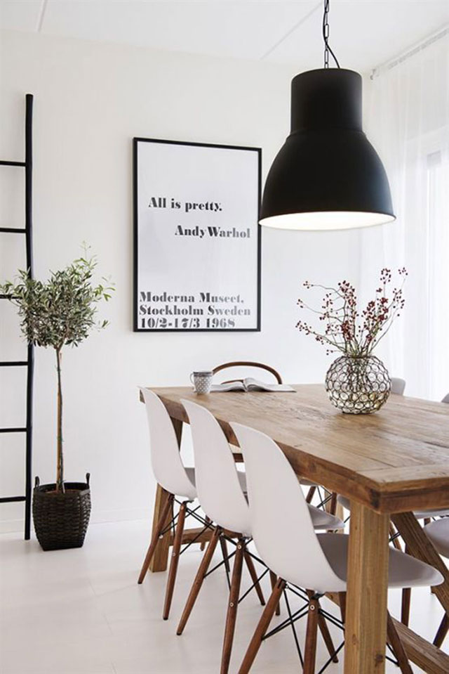 Gran mesa de comedor de madera natural con sillas blancas de patas de madera. Sobre la mesa una lámpara de pantalla negra. Comedor de muro, cielo y piso blancos.