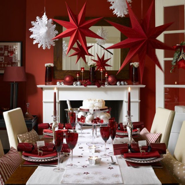 Comedor con mesa de mantel blanco, menaje en blanco con rojo, servilletas, copas y velas rojas. Del techo cuelgan estrellas grandes de cartón rojo y copos de nieve en papel blanco.