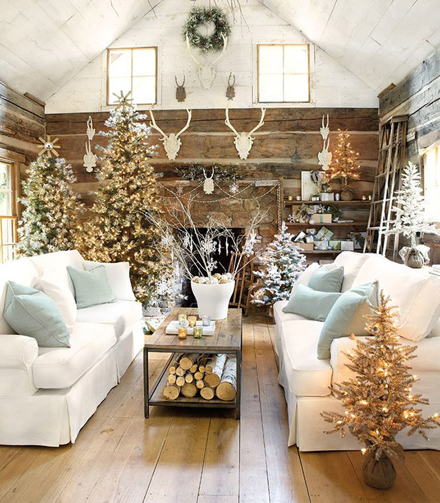 Sala de estar decorada en estilo navideño rústico o forest, con varios pinos de distintos tamaños decorados solo con luces. Hay pinos nevados, otros más verdes y otros en tonos ocres. Sillones de tela blancos con cojines celestes y al centro una mesa con leña, sobre la mesa un gran macetero blanco con ramas. Los muros, de tablones de madera, están decorados con cabezas de reno blancas.