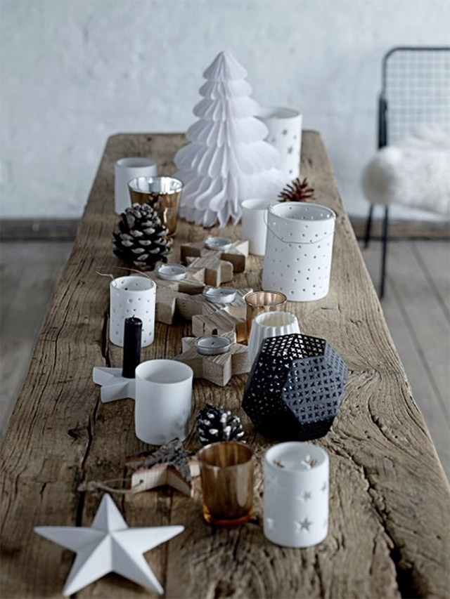 Mesa hecha de un tablón de madera en bruto decorada con estrellas, porta velas en forma de estrella, farolitos y pino de papel blancos. Además piñas de pino, porta velas de estrellas de madera, vasos dorados y porta vela hexagonal negro.