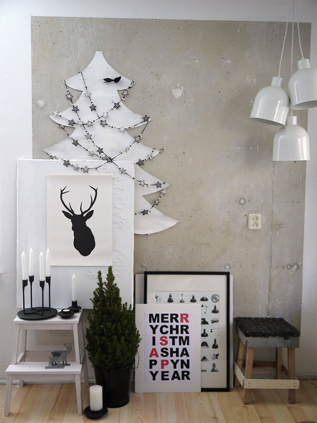 Muro de concreto a la vista decorado con la forma de un pino en blanco del que cuelgan guirnaldas con estrellas negras y blancas. Sobre una banqueta de madera blanca, un candelabro negro con velas blancas y apollándose en el muro un cuadro blanco con la silueta de un reno en negro. Dentro de un balde de lata negro un pino pequeño.