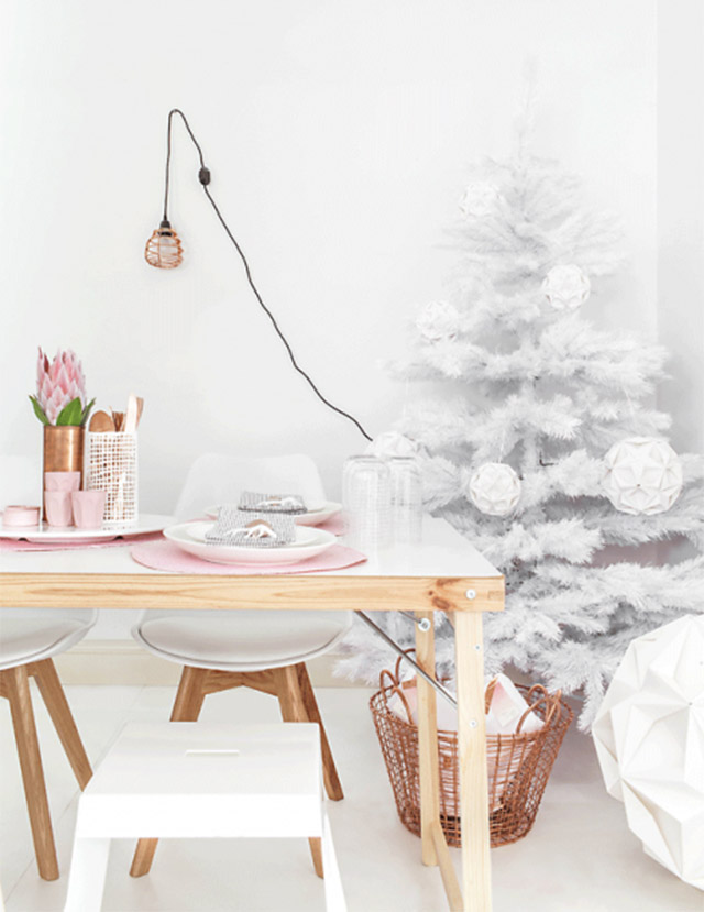 Mesa de madera con superficie lacada blanca, sillas blancas y patas de madera. Sobre la mesa, individuales rosados, vasos en el mismo color, florero de cobre con flores rosadas, un porta cubiertos calado blanco con cubiertos de madera. A un costado, canasto de fibra natural con tejido abierto en color cobrizo. Al fondo, pino de navidad blanco con adornos de esferas blancas.