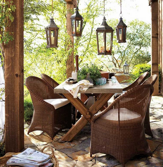 Juego de comedor de terraza con mesa de madera y sillones de mimbre con cojines blancos en los asientos. Del techo cuelgan unos faroles portavelas.