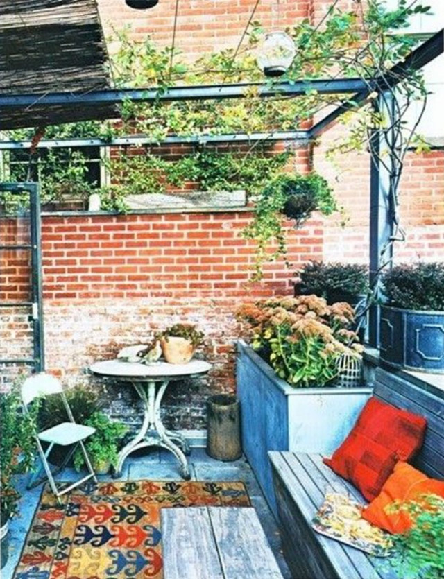 Terraza con mesa redonda y una silla plegable blanca. Alfombra de fondo amarillo y motivos de colores azul, rojo y verde. Jardinera alta con flores tipo hortensias. Banqueta de madera con cojines de colores rojo y anaranjado.