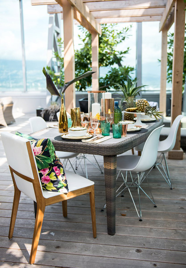 Comedor para terraza con mesa de ratán, sillas blancas de patas metálicas y sillas blancas estructura de madera, la silla de cabecera contrasta con un cojín negro con estampado floral de colores bien intensos. Mesa puesta con menaje en colores verde, blanco, aplicaciones doradas y cobre.