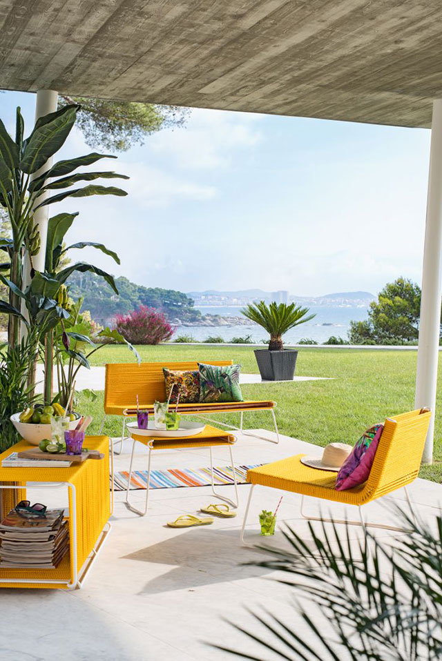 Juego de living de terraza en color amarillo, un sillón y una silla, mesa de centro y mueble. Cojines con diseño estilo tropical en colores verdes, fucsias y amarillo, vasos en colores fucsia, verde y transparente.