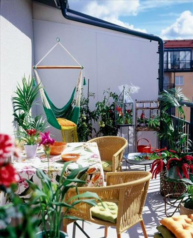 Terraza con juego de comedor de sillas de ratán amarillo y cojines verdes, mantel de la mesa blanco con estampado de hibiscus, hamaca verde tipo silla colgando de una esquina de la terraza. Maceteros con palmeras y plantas con flores grandes.