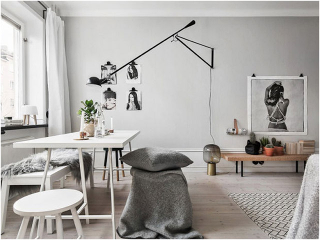 Sala de estar y comedor en una sola habitación. Muros de color gris claro, cielo blanco y piso de madera pálido. La decoración es en blancos como la cortina de la ventana, la mesa, piso y banqueta. Negro en repisa, lámpara y pisos. Madera y mucha textura natural, como pieles y mantas de lana. Las imágenes de la pared son en blanco y negro, sin marcos, sujetas con clips negros.