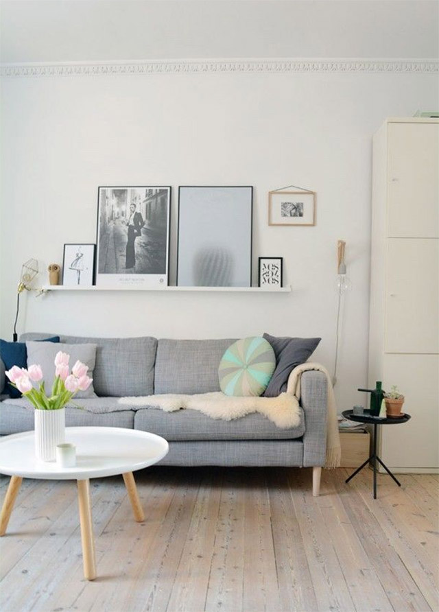 Sala de estar, muros y cielo blancos, piso de madera natural. Mesa de centro redonda de cubierta blanca y patas de madera, un sofá de tela gris. Cojines en colores azul, celeste, verde, además de manta de piel blanca sobre el sofá.