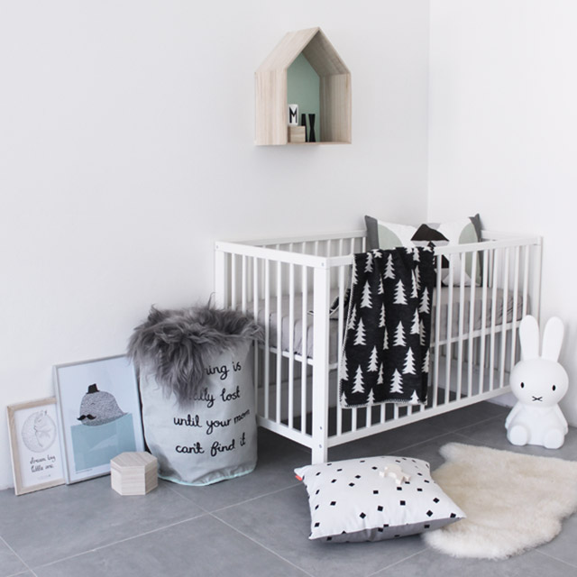 Dormitorio infantil, con cuna de madera blanca. Cojín dentro de la cuna de figuras geométricas en colores gris, blanco, negro y verde claro. Sobre el suelo bajada de cama de piel blanca y cojín blanco con pequeños cuadros negros.