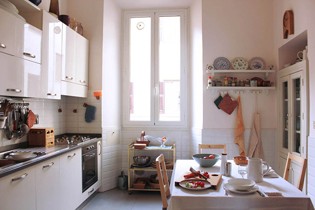 Cocina y comedor en tonos blancos. Muros mitad pintados y mitad cerámica, muebles de cocina blancos, mesa de comedor con mantel blanco y sillas de madera.