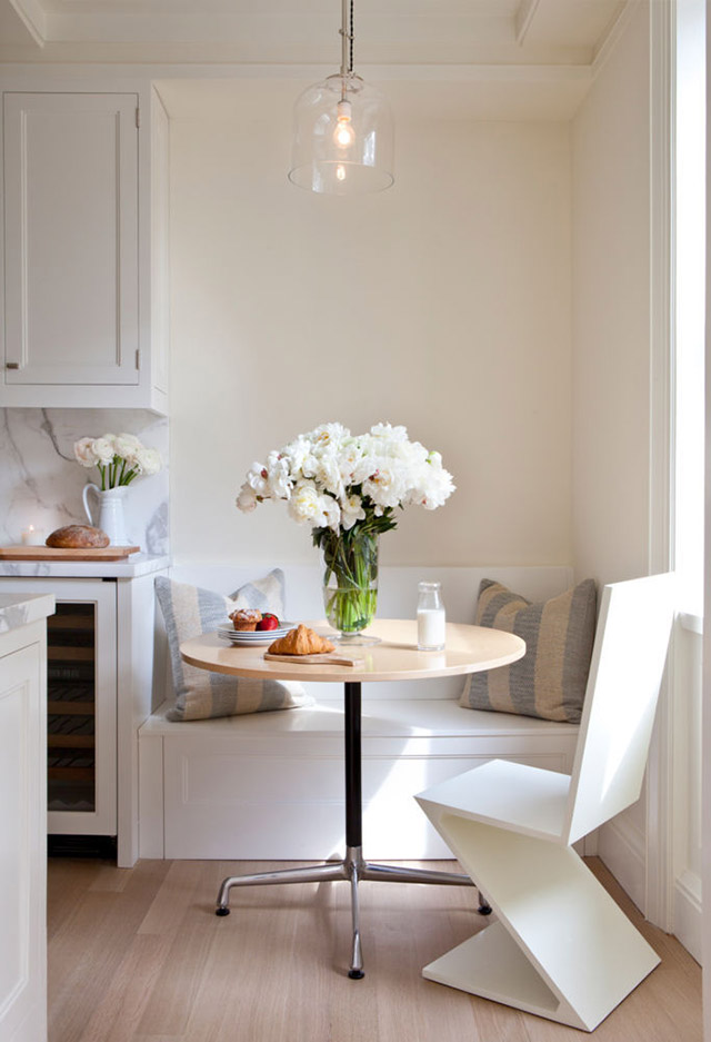 Adosada al muro una pequeña banqueta como continuación de la cocina, en madera laminada blanca, dos cojines en tonos celeste y crema, una mesa redonda de patas metálicas y silla de madera blanca de una sola pieza con patas en Z.