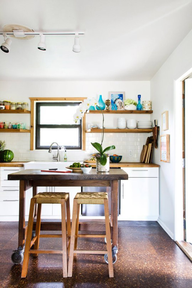 Cocina abierta con muebles de puertas blancas y cubierta de madera color natural, marco de ventana y repisas de madera color natural. Al centro de la cocina una mesa alta cuadrada de madera y ruedas en las patas, dos pisos de madera con asiento tejido en fibra natural.