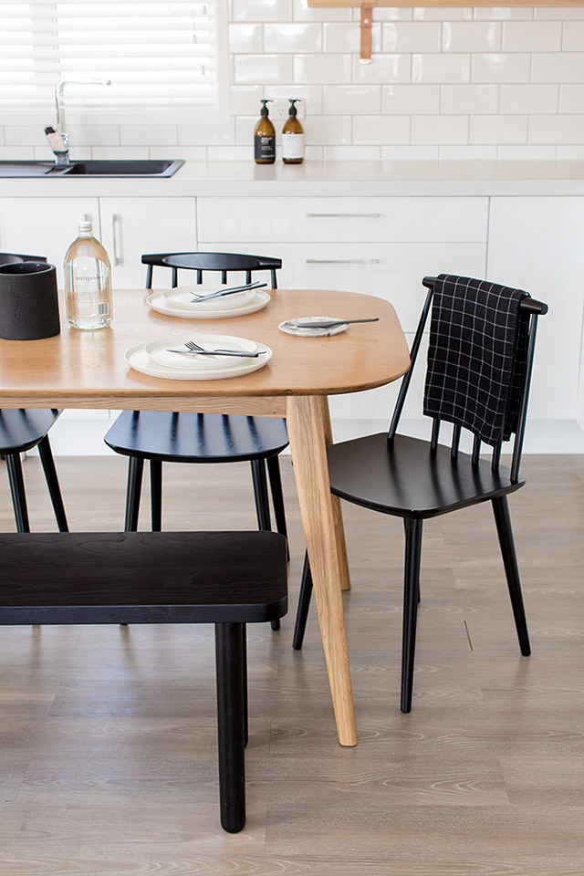 Mesa de comedor de madera estilo mid-century en la cocina de muebles y muros de cerámica blancos. Las sillas y banqueta junto a la mesa son de madera pintadas negro.