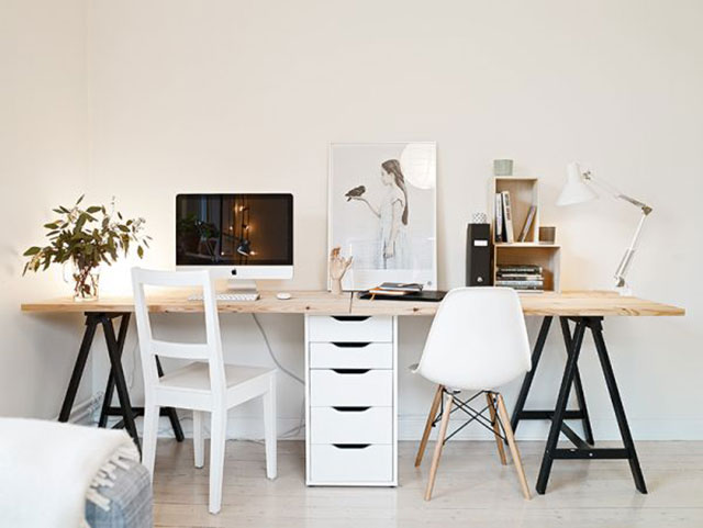 Mesón de escritorio de cubierta de madera color natural y base metálica negra para dos puestos de trabajo, separados por una cajonera blanca. Cada puesto con una silla blanca de estilos diferentes. Adornos y articulos de organización sobre el escritorio. Muros blancos sin decoración.