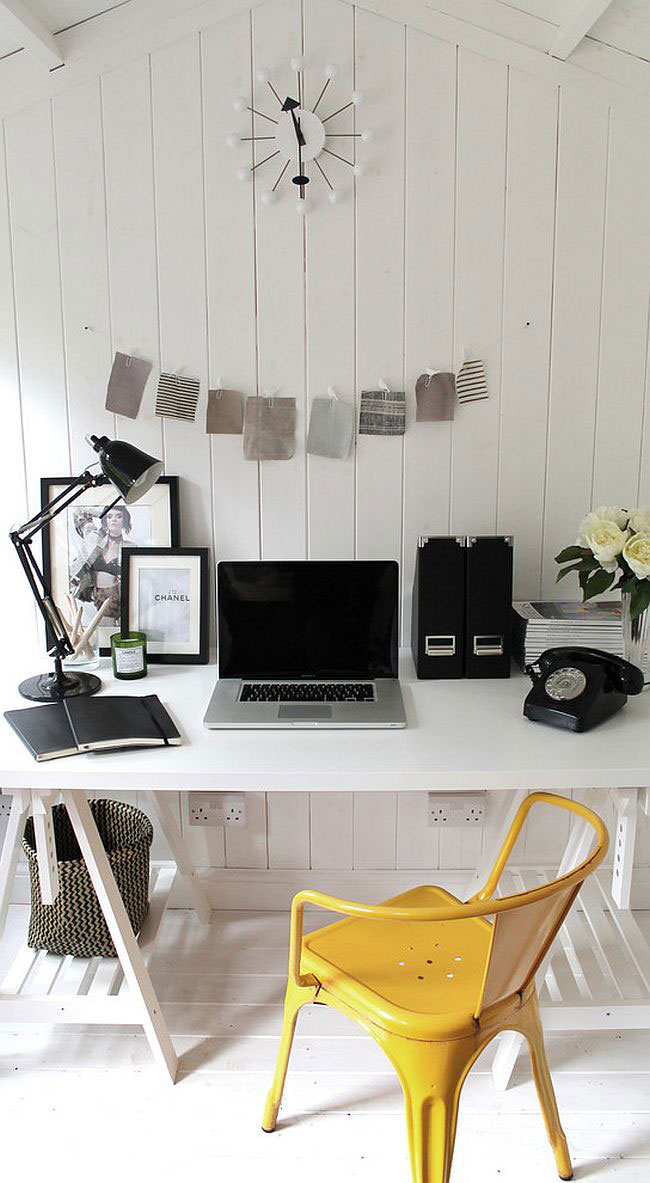 Espacio de trabajo con piso, muro y cielo de madera pintados blancos. El escritorio es de madera pintada en blanco con patas de atril. Silla metálica amarilla. Decoración sobre el escritorio en blanco y negro. Sobre el muro, una muestra de retazos de tela en tonos grises y un reloj blanco con el minutero y segundero negros.