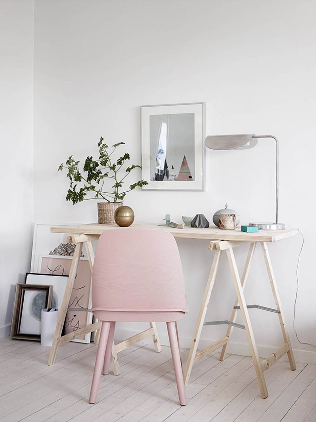 Escritorio de madera color natural, patas de atril. Silla de madera en rosa cuarzo. Decoración con cuadros apoyados en el piso y la pared, solo un cuadro colgado al muro frente al escritorio. Simple y muy femenino.