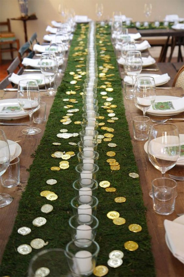 Una gran mesa servida para celebrar San Patricio. El camino de mesa son palmetas de pasto con velas blancas y esparcidas alrededor moneditas de oro de chocolate.