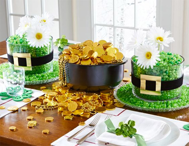 Mesa celebrando San Patricio. Individuales y loza blancos, las servilletas con adornos de cinta verde y tréboles, los posavasos son tréboles y, el centro de mesa es una gran olla con monedas de chocolate de oro y dos sombreros de duende con caramelos verdes. Los sombreros adornados con margaritas