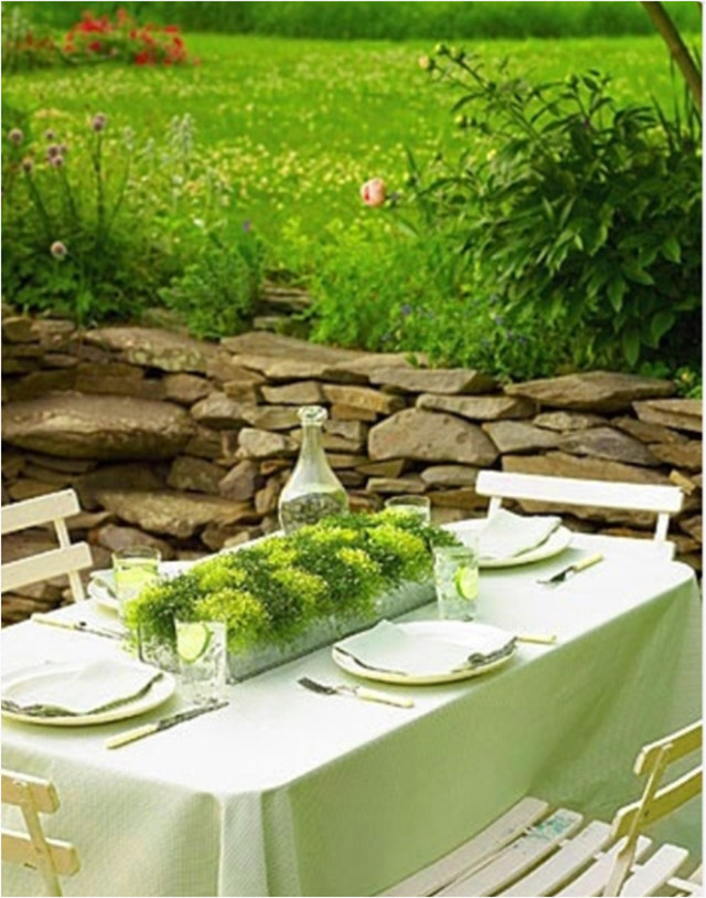 Mesa al aire libre con mantel verde claro, centro de mesa con pequeñas plantas en distintos tonos de verde.